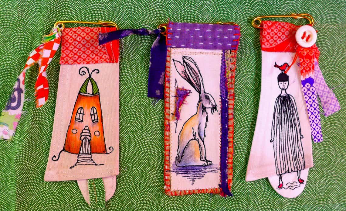 Three decorative fabric brooches featuring unique designs: a whimsical house, a rabbit, and a figure in a dress, displayed against a green background.